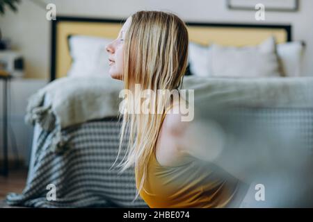 Giovane donna con lunghi capelli biondi in giallo sport top fa esercizi vicino al letto coperto con coperta a righe con cuscini in cima Foto Stock