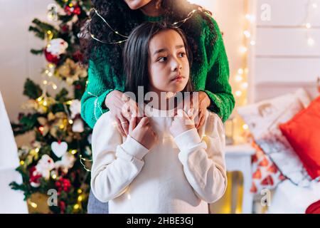 Ritratto di una bambina e sua madre in piedi dietro di lei in un maglione verde, mettendo le mani sulle sue spalle sullo sfondo di un albero di Natale con una ghirlanda, una ragazza e mamma stanno tenendo le mani Foto Stock