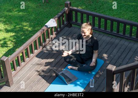 una donna sta facendo yoga sul portico della sua casa sorridendo meditazione nella posizione del loto. c'è un computer accanto ad esso classi online con un maestro Foto Stock