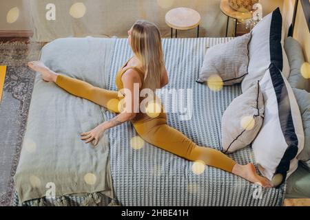 Donna atletica con lunghi capelli biondi in stretching sportivo facendo split su soffice coperta letto con cuscini e illuminazione ghirlanda Foto Stock