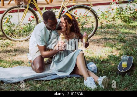 Il giovane uomo afro-americano e donna bruna si siedono sul copriletto su erba mangiando gelato e ridendo nella giornata estiva di sole nel parco Foto Stock