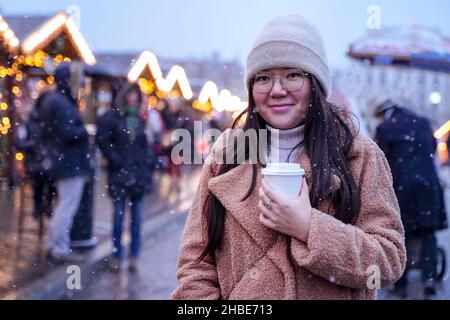 Ragazza carina sullo sfondo della città di Natale in inverno. Giovane donna asiatica che si gode a piedi e fiera in tempo nevoso Foto Stock