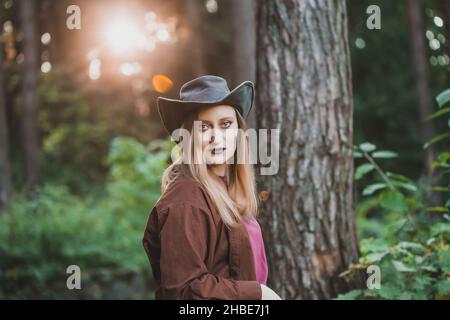 Ritratto di moda in stile country di una donna bionda che indossa un cappello da cowboy in pelle e una camicia marrone in piedi nella foresta. Drammatico pesante make up con scuro Foto Stock