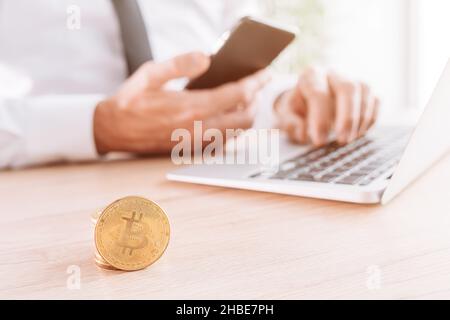 Businessman che commercia con la piattaforma di commercio di criptovaluta del bitcoin sopra il suo smartphone ed il calcolatore del laptop in ufficio, monete impilate davanti, selettive Foto Stock