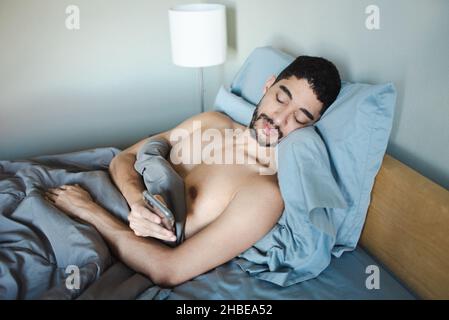 L'uomo milleniale bearded di razza mista propped su cuscini che controllano il telefono nel letto la mattina mentre si sveglia. Letto con lenzuola blu e trapunta blu Foto Stock