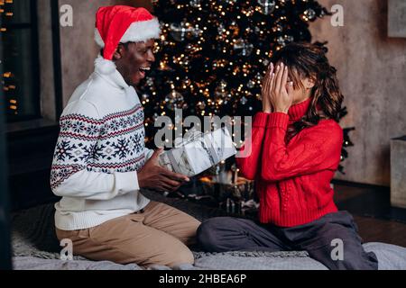 L'uomo afro-americano in un cappello rosso dà un regalo di Natale una scatola sullo sfondo di un albero di Capodanno, una coppia in amore celebra a casa, una donna in un maglione rosso copre il suo volto con le mani ed è in attesa di una sorpresa Foto Stock