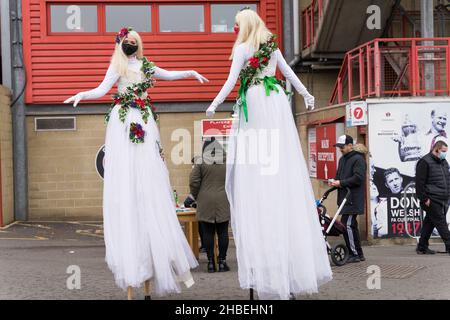 Greenwich London UK 19 dicembre 2021. Il gruppo Southeast London Clinical Commissioning si è accollata questo fine settimana la vaccinazione covid-19 per tutti gli adulti idonei al The Valley, Charlton Athletic FC. Due giovani donne vestite su palafitte che si esibiscono all'ingresso del centro di vaccinazione accolgono le persone per essere vaccinate per fermare l'impennata della variante Omicron del coronavirus in inverno che conduce alla stagione natalizia. Credit: Xiu Bao/Alamy Live News Foto Stock