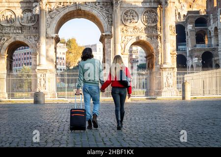Giovane coppia in viaggio a Roma. La giovane coppia cammina e tiene le mani. L'uomo sta trascinando il suo carrello. Foto Stock