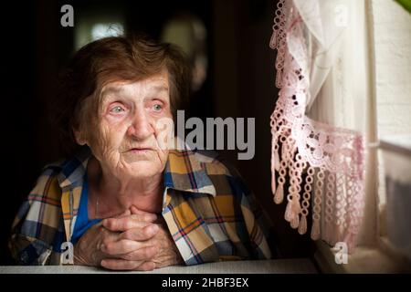 Una donna anziana siede pensierosamente alla finestra. Foto Stock
