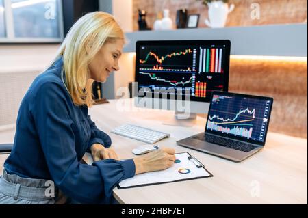Investitore e Crypto trader donna matura e intelligente di successo, utilizzando pc e laptop, analizza grafici di trading in borsa e lo scambio di criptovaluta digitale, conduce analisi, trading cripto monete Foto Stock
