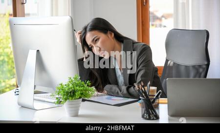 Una donna d'affari stanca si siede alla sua scrivania in ufficio, lavorando su un computer desktop.A giovane donna asiatica si sente sopraffatta, esausta e annoiata Foto Stock