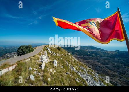 Sulla cima del monte Lovcen, la maestosa bandiera nazionale rossa dello stato balcanico che sbatte nel vento, contro cieli limpidi e soleggiati blu di fine estate Foto Stock