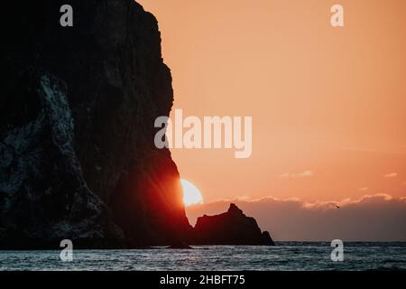 Un tramonto rosso acceso sul mare con scogliera vulcanica rocciosa. Astratta natura estate o primavera oceano mare sfondo. Piccole onde su acqua calda dorata Foto Stock