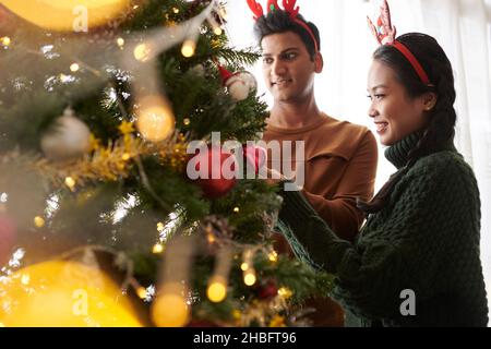 Giovane marito e moglie che decorano l'albero di Natale a casa per festeggiare Foto Stock