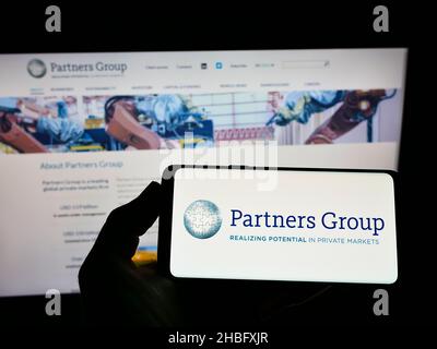 Persona che tiene uno smartphone con il logo della società di investimento svizzera Partners Group Holding AG sullo schermo di fronte al sito web. Mettere a fuoco sul display del telefono. Foto Stock