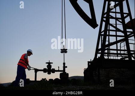 Operatore petrolifero in gilet da lavoro che utilizza la macchina a bilanciere della pompa del petrolio per estrarre il petrolio grezzo dal pozzo. Operatore maschio che aziona il martinetto della pompa del petrolio durante il lavoro in campo petrolifero. Concetto di estrazione dell'olio. Foto Stock