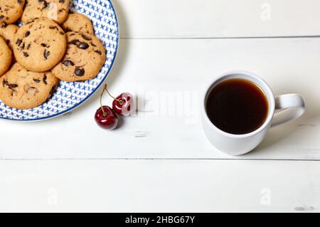 Tazza bianca con caffè, biscotti tradizionali e ciliegie dolci su sfondo bianco di legno, primo piano. Colazione gustosa, concetto di routine mattutino Foto Stock