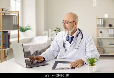 Medico senior in occhiali e abito medico bianco che lavora su un computer portatile seduto sul posto di lavoro in ufficio. Foto Stock