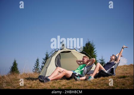 Donna escursionista che si allunga e sorride mentre si siede accanto al marito vicino tenda turistica. Coppia felice viaggiatori che riposano mentre camminano insieme in montagna. Concetto di escursioni, campeggio e relazioni. Foto Stock