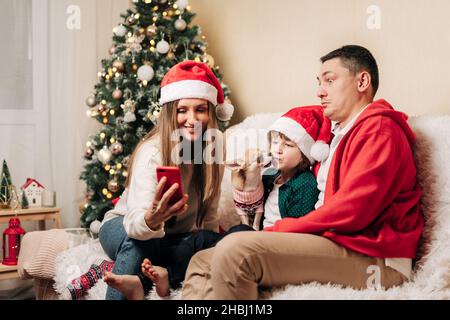 Famiglia felice con figlio bambino che ha una video chat al telefono. Madre in Santa Hat, padre, bambino ragazzo e cane in maglione che si diverte durante le vacanze di Natale a. Foto Stock