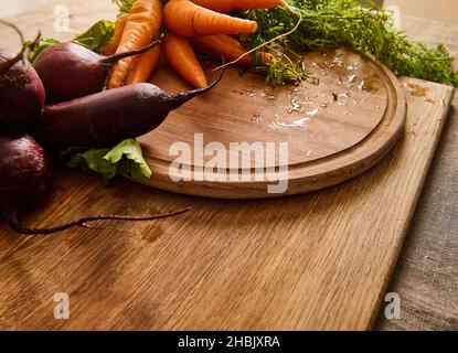 Mazzi di barbabietole fresche e biologiche e carote per bambini con cime su un tagliere di legno. Sfondo vegano cibo. Verdure crude di stagione. Foto Stock