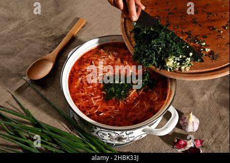 Vista dall'alto delle mani dello chef riempire il borscht appena bollito con aneto fresco tritato, erbe e aglio. Composizione alimentare con pentola di sou tradizionale ucraino Foto Stock