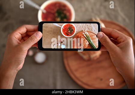 Vista dall'alto delle mani che tengono lo smartphone e fotografano una ciotola con zuppa tradizionale Ucraina appena cucinata - Borscht con cipolla fresca cruda verde Foto Stock