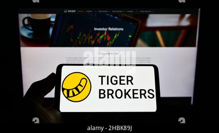 Persona che detiene il cellulare con il logo della società UP Fintech Holding Limited (Tiger Brokers) sullo schermo di fronte alla pagina web. Mettere a fuoco sul display del telefono. Foto Stock