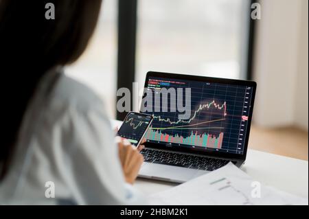 Una donna intelligente di successo Crypto trader, un investitore, utilizzando computer portatile e telefono cellulare, analizza le carte di trading nel mercato azionario di scambio di criptovaluta digitale, analizza, acquista e vende cripto monete Foto Stock