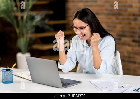 Sorpresi successo sicuro emozionale caucasica donna d'affari, broker, manager indossare occhiali, seduto a tavola con laptop, godendo di grande profitto, vittoria, buone notizie, gesturing con pugni, sorridente Foto Stock