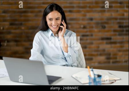 Bruna caucasica bruna donna di successo carina, SPECIALISTA IT, manager, seduto alla scrivania al computer portatile, parlare sullo smartphone mentre si lavora con colleghi o clienti, sorridente amichevole Foto Stock