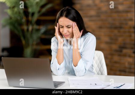Caucasica giovane adulta donna d'affari, manager o segretario che sperimenta stress sul lavoro in ufficio, stanco di sedersi al lavoro su un computer portatile, massaggiando templi, chiudere gli occhi, ha bisogno di riposo Foto Stock
