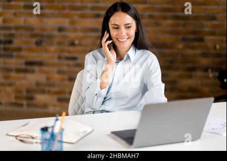 Allegro bruna caucasica amichevole, donna d'affari, specialista IT, manager, seduto alla scrivania al computer portatile, parlare al telefono mentre si lavora con amici o clienti, sorridente amichevole, guardando lo schermo Foto Stock