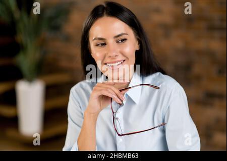 Bella bruna giovane adulto bruna caucasica successo, amministratore delegato o broker indossare occhiali, seduto in ufficio, guardando attentamente da parte, sognando di vacanza, sorridente Foto Stock