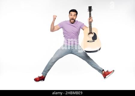 uomo sorpreso in jeans blu e t-shirt viola levitating tenendo la chitarra acustica su bianco Foto Stock