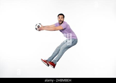 uomo emozionato in jeans e t-shirt viola che si levitating con il calcio su bianco Foto Stock