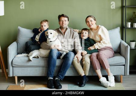 Ritratto completo di grande famiglia felice con due bambini e cane seduto sul divano a casa contro il muro verde, spazio copia Foto Stock