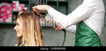 Creare ricci con i ferri da stiro. Parrucchiere fa un acconciatura per una giovane donna con lunghi capelli rossi in un salone di bellezza Foto Stock