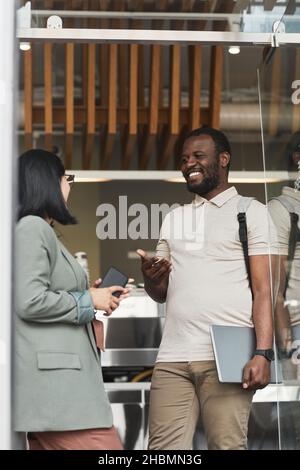 Ritratto verticale di un uomo afroamericano sorridente che parla con la giovane donna mentre si alza in piedi nella porta del caffè Foto Stock