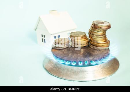 Fiamme blu che circondano pile di monete su un fornello a gas, con una casa bianca in miniatura sullo sfondo. Concetto creativo che rappresenta i prezzi del gas Foto Stock