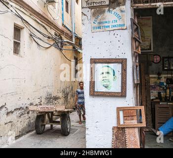 CITTÀ DI PIETRA, ZANZIBAR, TANZANIA - 13 MARZO 2017: Strada stretta in Stone Town con un giovane uomo, un carrello di legno e un negozio di cornici Foto Stock