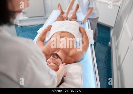 Il terapista massaggia la testa dell'uomo mentre il giovane collega lavora con le gambe in ospedale Foto Stock