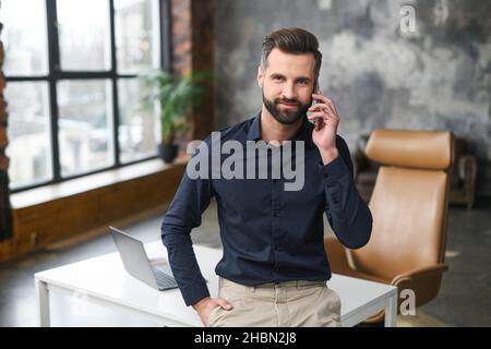 Felice ispirato caucasico maschio piccola impresa proprietario parlare sullo smartphone mentre in piedi in un moderno ufficio loft. Sorridente uomo manager contemporaneo di successo in abbigliamento casual tenendo una telefonata Foto Stock