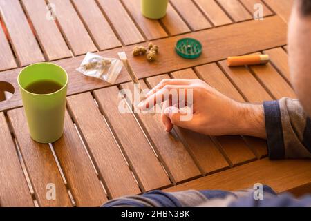 Giovane uomo che fuma una marijuana comune con una bevanda in un luogo all'aperto durante una giornata di sole. Foto Stock