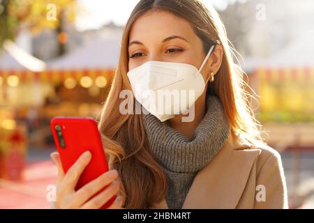 Primo piano di bella ragazza con maschera medica FFP2 KN95 guardando il suo smartphone nei mercati di Natale Foto Stock