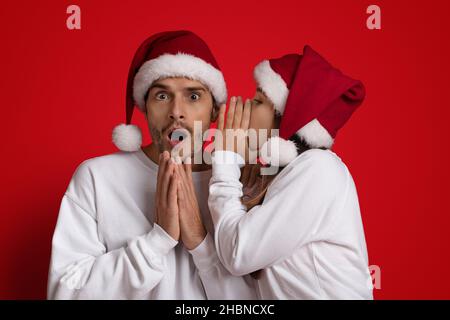 Giovane donna che indossa Santa Hat Sharing Secret con il suo ragazzo scioccato Foto Stock