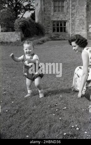 1950s, storico, fuori, nel terreno di una casa di campagna, un ragazzino felice, essendo stato liberato dal contratto d'affitto, vale a dire, la sua imbracatura da sua madre, è eccitato per essere sui suoi due piedi, Inghilterra, Regno Unito. Foto Stock