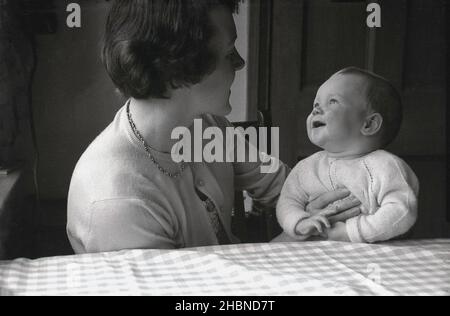 1950s, storico, ad un tavolo da cucina, una madre, seduta tenendo e guardando il suo giovane figlio neonato, che la guarda indietro, sorridente, Inghilterra, Regno Unito. Foto Stock