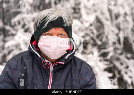 Anziana donna anziana con capelli grigi indossando rosa usa e getta virus maschera, sfocato inverno neve coperto alberi sfondo Foto Stock