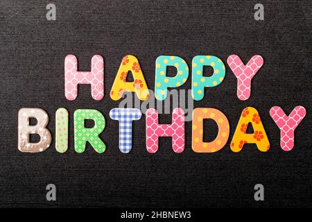 Carta con Happy Birthday parole fatte da lettere di legno colorate e vivide miste su un materiale tessile nero scuro testurizzato che può essere utilizzato come messaggio Foto Stock